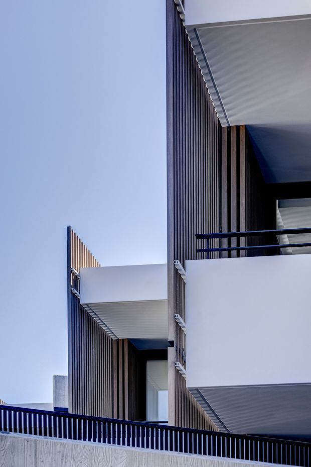 Vertical architectural photograph of a contemporary multi-storey building facade, featuring projecting white balcony slabs and canopies, dark vertical timber-like screening, black metal balustrades, and strong geometric shadow lines against a clear sky.