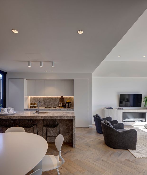 Open-plan contemporary kitchen, dining and living area with a compact island, integrated pale cabinetry, stone benchtop and warm illuminated backsplash. The living zone includes curved dark upholstered lounge chairs, a wall-mounted television and low built-in media cabinetry. Light herringbone timber flooring, recessed lighting and a neutral palette create a refined, modern interior.
