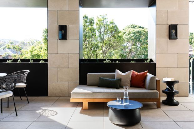A bright, contemporary covered outdoor lounge or terrace with large open glazed openings framing trees and neighbouring rooftops. The space features a low upholstered sofa with contrasting cushions, a round black coffee table, sculptural side table, woven dining chairs, tiled flooring, stone-look tiled columns, dark balustrade panels, and integrated wall lights. The setting has a refined indoor-outdoor character with strong natural light and views to the surrounding landscape.
