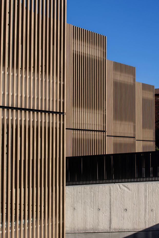 Exterior architectural view of a contemporary multi-level facade composed of closely spaced vertical timber battens, creating a screened and rhythmically articulated envelope. Black metal railings and trim contrast with pale textured concrete or cement-board surfaces at the lower level. A small portion of an adjacent brick building is visible at the right, beneath a clear blue sky.