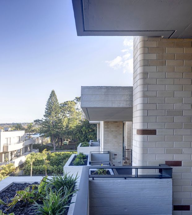 Exterior view from a modern apartment balcony or terrace, looking along a multi-level building with pale rendered brick walls, projecting concrete balcony slabs, dark metal balustrades, and raised planter beds with landscaping. The elevated setting overlooks neighbouring homes and mature trees under a bright sky.