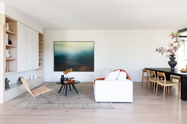 Bright open-plan contemporary living and dining area with white built-in cabinetry and shelving, pale timber flooring, a large neutral rug, white sofa, sculptural coffee table, timber lounge chair, and a dark dining table with matching timber chairs. A large abstract artwork and decorative vase add restrained visual accents. The space has a clean, calm, refined appearance with abundant natural light.