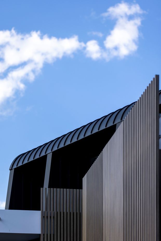 A cropped architectural exterior detail against a bright blue sky, showing a contemporary building with a dramatic curved ribbed roof edge, tall vertical metal or aluminium screening, dark recessed openings, and a white lower wall. The composition emphasizes strong diagonal lines, shadow, and sculptural facade geometry.