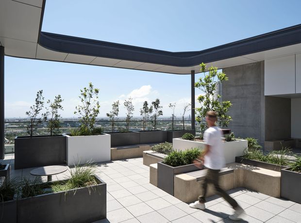 A contemporary rooftop terrace with expansive city views, framed by a dramatic curved roof overhang. The terrace features large-format tiled paving, integrated concrete and dark metal planter boxes, built-in bench seating, sculptural greenery, and a glass balustrade. A modern building interior is partially visible at right, while a blurred person walks through the space.