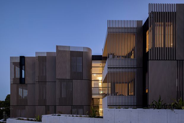 Twilight exterior view of a contemporary multi-storey building with strongly articulated vertical slatted screens, dark cladding, recessed illuminated glazing, projecting volumes, and exposed concrete retaining walls. The layered facade creates a sculptural, privacy-screened composition.