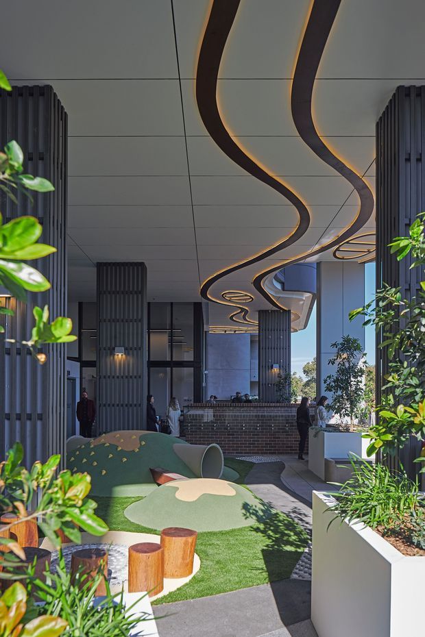 A landscaped open-air courtyard or entrance plaza beneath a large covered canopy. The ceiling features sweeping, ribbon-like dark panels with warm concealed lighting, set against a pale panelled soffit. Tall dark ribbed architectural columns frame glazed building fronts and a brick reception or service counter in the background. Lush planters, artificial or manicured turf, sculptural curved green seating, and small timber stools create a contemporary, inviting communal space.