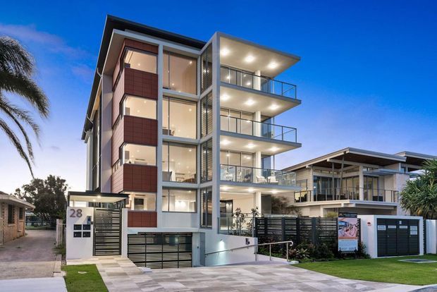 Twilight exterior view of a contemporary multi-storey residential apartment building with strong vertical glazing, projecting balconies, glass balustrades, dark metal framing, and pale rendered facade elements. A lower neighbouring or attached dwelling with a broad roof overhang is visible to the right, along with landscaped frontage, driveway paving, entry gates, and palm trees.