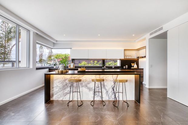 Bright, spacious contemporary kitchen with a large island featuring a dramatic marble-look waterfall benchtop and dark cabinetry. Three bar stools sit at the island, with extensive white built-in cabinetry, integrated appliances, open shelving, and large windows providing abundant natural light. Dark stone-look flooring and black accents create a refined, high-contrast interior.