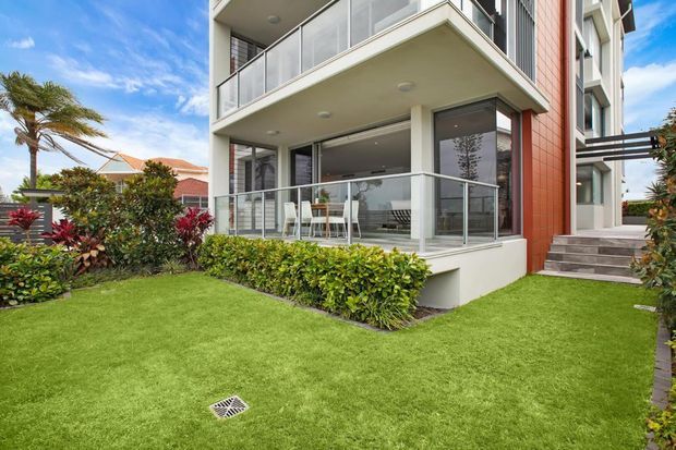 Exterior view of a contemporary multi-storey residential building with stacked balconies, clear glass balustrades, pale rendered walls, and a prominent warm reddish-brown vertical cladding panel. The ground-floor balcony opens onto a manicured lawn with landscaped planting, while broad glazing reveals a light interior dining or living area. Adjacent homes and tropical landscaping are visible in the background.