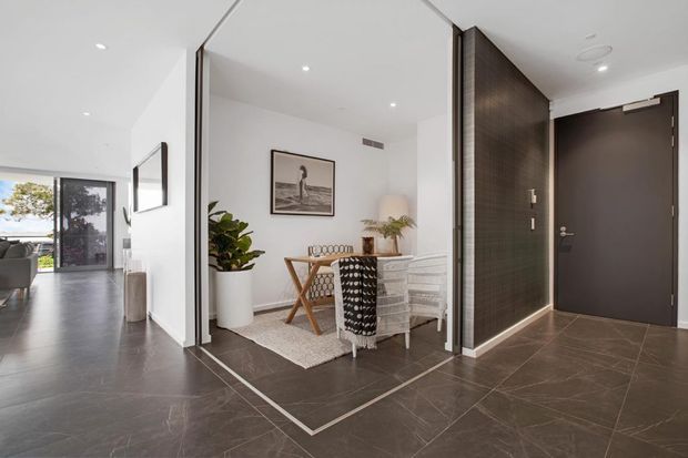 Bright contemporary apartment entry and dining area with dark large-format tiled flooring, white walls, a dark timber-look feature partition, and a compact dining table with chairs. The open-plan living area and exterior glazing are visible to the left.