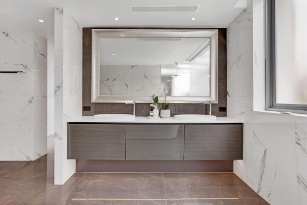 A refined contemporary bathroom featuring a wide floating timber-toned vanity with integrated basins, a large framed mirror, white marble-look wall panels with grey veining, and warm brown stone-look flooring. The space has recessed ceiling lighting, a wall-mounted split-system unit, and a window providing natural light.