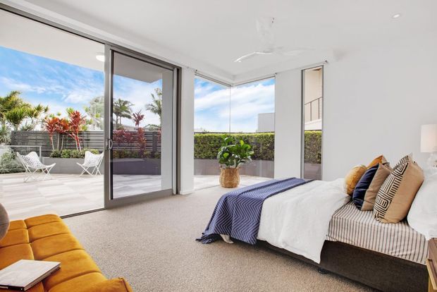 Bright contemporary bedroom with a low upholstered bed, layered neutral and patterned bedding, and a mustard tufted bench. Large sliding glass doors open to a private terrace or balcony with outdoor chairs, planter boxes, and tropical landscaping. The room has white walls, beige carpet, slim dark window frames, and abundant natural light.