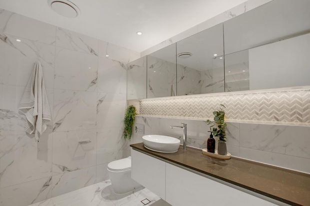 Bright contemporary bathroom featuring large white marble-look wall and floor tiles, a floating white vanity with a dark stone benchtop, vessel basin, wall-to-wall mirrored cabinetry, and a decorative chevron mosaic feature strip with concealed lighting. A toilet and shower area are visible.