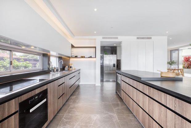Contemporary galley-style kitchen with extensive dark timber cabinetry, black stone benchtops, a large central island, integrated appliances, and light grey stone-look flooring. White walls and ceiling, recessed lighting, and broad windows create a bright, clean space.