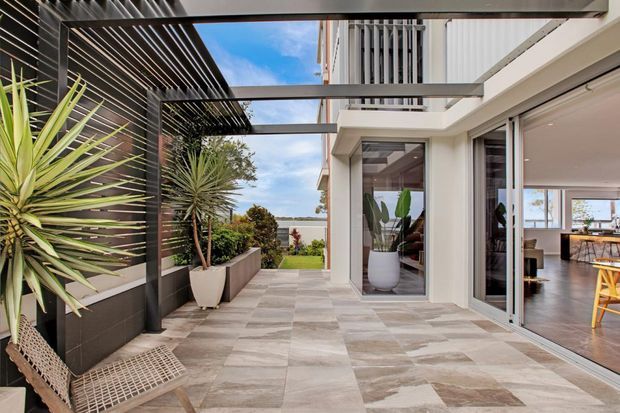 A contemporary covered outdoor terrace or courtyard beside a modern multi-level home. The space features large-format neutral stone-look paving, dark vertical wall cladding, a slatted pergola with exposed steel beams, architectural potted plants, and sliding glass doors opening into a bright dining area. The terrace overlooks a landscaped garden and distant water or sky views.