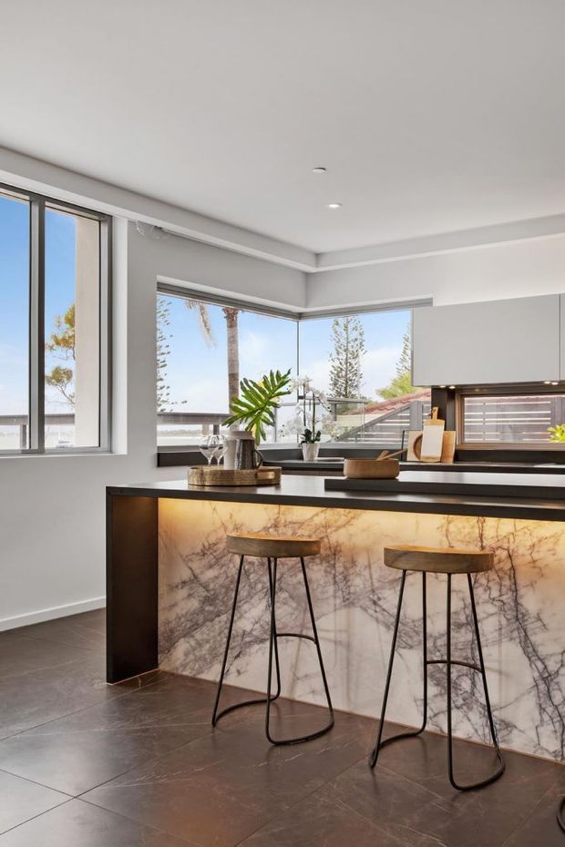 A contemporary kitchen interior with a large dark island featuring a dramatic veined stone waterfall-style front and warm under-counter lighting. Two slim timber-and-black metal bar stools sit at the island. White and grey cabinetry, dark tiled flooring, recessed lighting, and extensive corner glazing create a bright, refined space with views to palm trees and neighbouring rooftops.