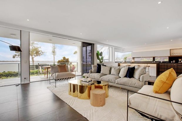 Bright, open-plan contemporary living area with a large sectional sofa, sculptural gold nesting coffee tables, a timber side table, and a cream armchair with a mustard cushion. The room has dark large-format flooring, a white ceiling with recessed downlights, extensive floor-to-ceiling glazing opening to a balcony or terrace, and a sleek kitchen with dark cabinetry and a light benchtop at the rear.