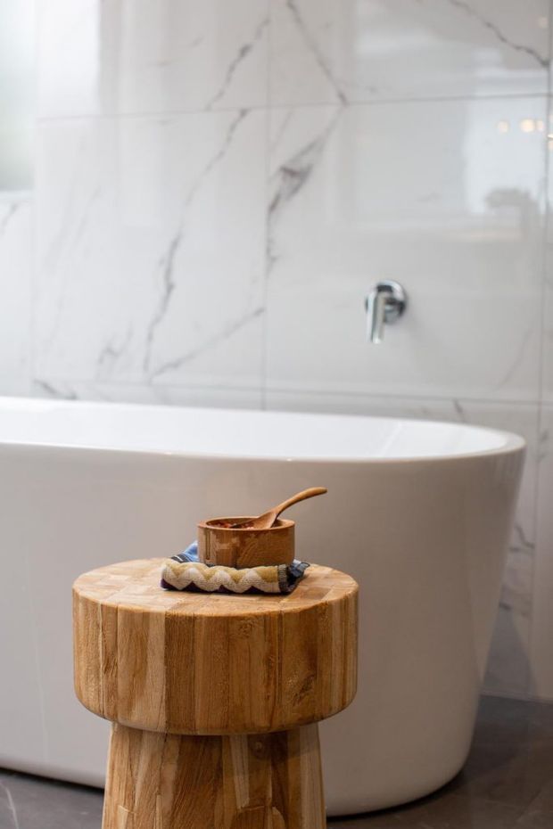 A refined bathroom scene featuring a freestanding white bathtub against large white marble-look wall tiles with subtle grey veining. A chrome wall-mounted tap is visible, while a round natural timber pedestal stool in the foreground holds a small wooden container and folded textile. The space has a bright, warm, spa-like atmosphere with dark flooring providing contrast.