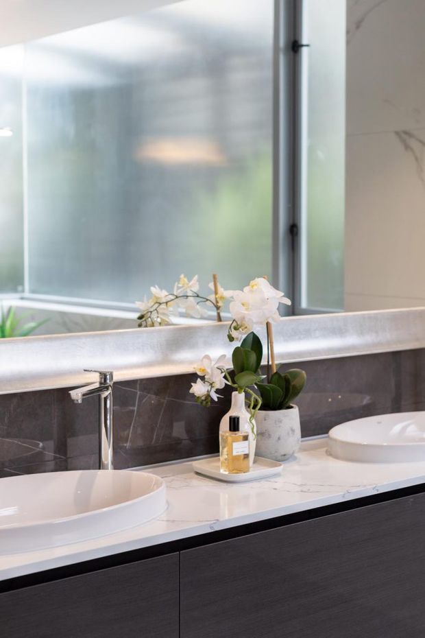 A refined modern bathroom vanity with a long white marble-look countertop, two white vessel basins, chrome tapware, and dark wood-grain cabinetry. A dark grey marble backsplash runs beneath a large mirror, with frosted glazing and an orchid adding softness.