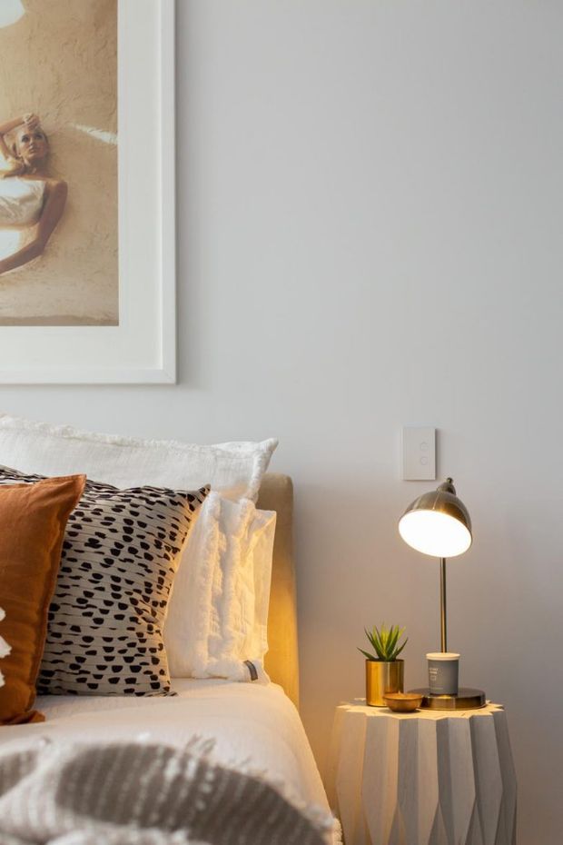Close-up view of a warmly styled bedroom corner featuring a partially visible bed with layered white bedding, a rust-orange cushion, patterned accent pillow and textured throw. A small sculptural side table holds a modern table lamp, potted plant and bowl, while framed artwork hangs on the pale grey wall above.