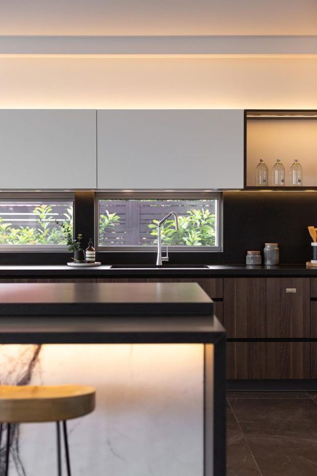 A refined contemporary kitchen featuring dark timber cabinetry, a black splashback and benchtop, pale grey overhead cupboards, integrated warm LED lighting, and a large island with a pale veined stone waterfall panel. Two horizontal windows overlook leafy planting, while a stainless steel sink and tap sit beneath the glazing. A timber stool is positioned at the island.