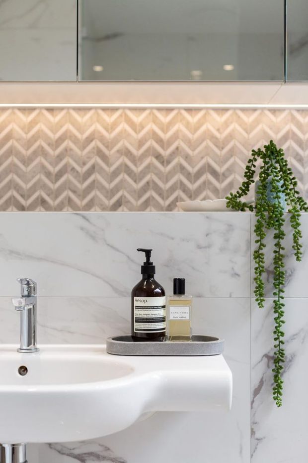 Close-up view of a refined bathroom vanity with a white wall-mounted basin, chrome tapware, large-format white marble-look wall tiles, and a decorative beige chevron-patterned tile splashback. Mirrored cabinetry is mounted above, with a trailing plant and neatly arranged soap dispensers on a grey stone tray. The overall look is polished, warm, and contemporary.