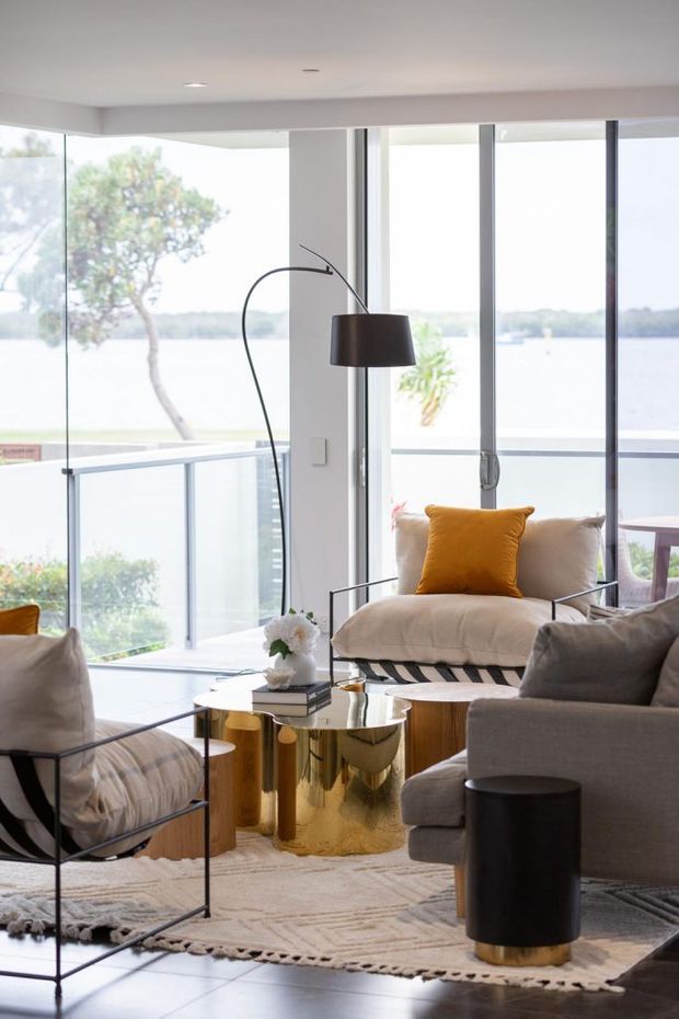 Bright contemporary living room overlooking a body of water through extensive floor-to-ceiling glazing. The room features upholstered lounge chairs and sofa seating, a sculptural black floor lamp, a reflective gold cylindrical coffee table, patterned neutral rug, and restrained white and grey finishes with a mustard accent cushion. A balcony with glass balustrade is visible beyond the glazing.