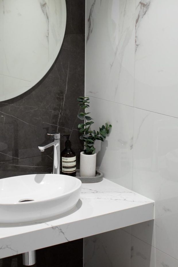 Close-up of a contemporary bathroom vanity featuring a white vessel basin, chrome faucet, white marble-look countertop and wall tiles, and a contrasting dark marble-look tile feature wall behind a round mirror. A small potted plant and soap dispenser sit on the vanity.