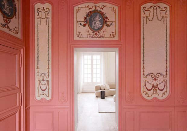 View through an ornate salmon-pink paneled doorway into a bright, sparsely furnished living room. The foreground walls and doors feature elaborate classical mouldings, painted decorative panels, and gilded ornamental motifs. The adjoining room has pale flooring, a light sofa, a small dark cylindrical table, and a large multi-pane window.