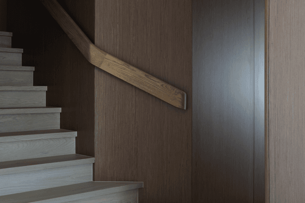 Close-up interior view of a contemporary stairway with pale timber treads and risers, a sculptural timber handrail mounted against a dark vertically textured wood wall, and a tall dark timber-clad wall or door panel. The composition is minimalist and moody, emphasizing the contrast between light and dark wood surfaces.