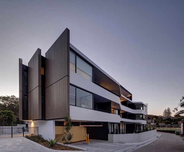 Contemporary multi-storey residential apartment building photographed from the street at dusk. The facade combines dark vertical fins, extensive glazing, white and charcoal projecting floor plates, curved balconies, recessed lighting, and a ground-level garage or service area. The building sits on a sloped urban/suburban site with landscaped edges and a driveway.