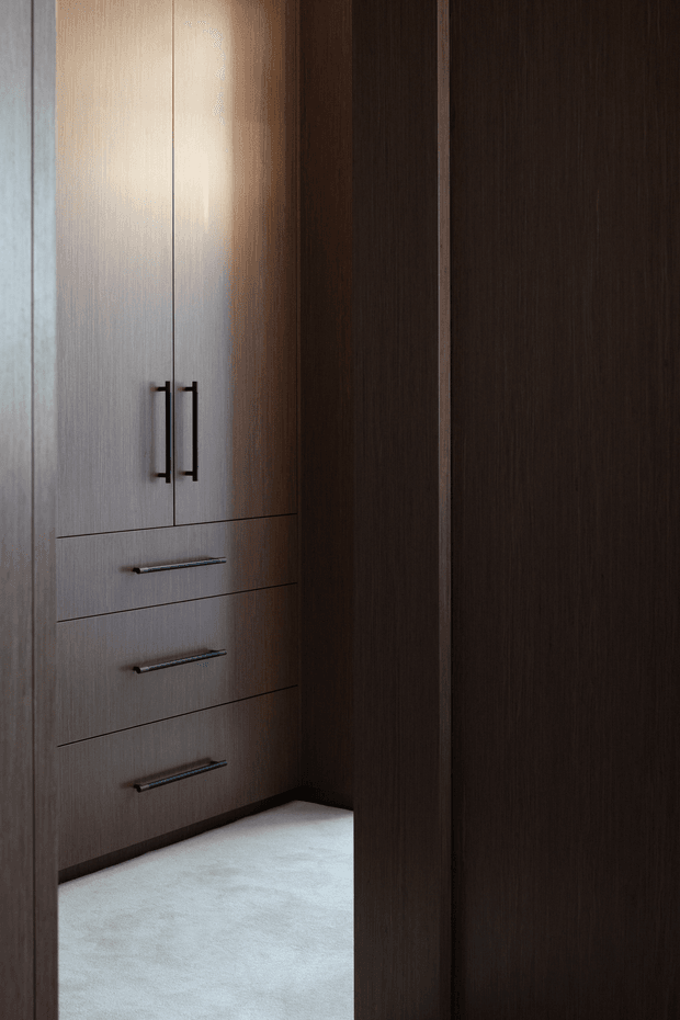A dark, refined walk-in wardrobe with floor-to-ceiling built-in timber cabinetry. The cabinetry includes tall double-door storage and three wide drawers with slim dark metal handles, contrasted against a pale carpeted floor. The space is narrow and softly lit, with warm wood tones and a minimalist, tailored appearance.