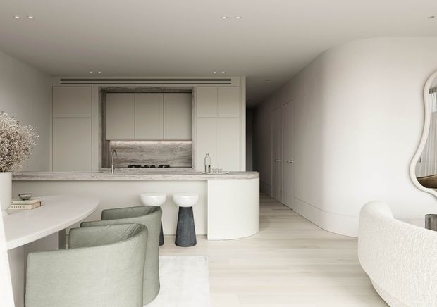 Bright, refined open-plan kitchen and dining area with a pale neutral palette. The kitchen features full-height white cabinetry, a stone benchtop and splashback, a sculptural curved island, and two stools. A round dining table with upholstered chairs occupies the foreground, while a softly curved boucle-style lounge chair and a wall mirror sit to the right. Recessed ceiling lighting, concealed appliances, and light timber flooring create a clean contemporary interior.