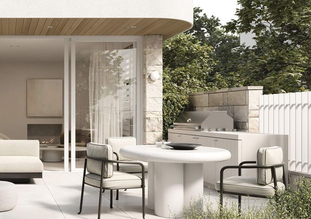 A refined outdoor dining and entertaining area adjoining a contemporary interior living space. The terrace features a rounded white dining table with upholstered black-framed chairs, a built-in outdoor kitchen with grill and cabinetry, pale stone walls, white fencing, extensive glazing, and a curved timber-lined soffit. The palette is light and neutral with strong indoor-outdoor connection and mature greenery beyond.