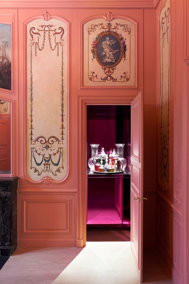 A richly decorated coral-pink interior with ornate raised wall panelling, framed decorative classical artwork, and detailed painted mouldings. An open concealed bar or drinks cabinet is built into the wall, illuminated in vivid magenta and displaying crystal decanters and glassware. The room has a polished dark wood floor, a pale area rug or light flooring in the foreground, and highly ornamental traditional detailing throughout.