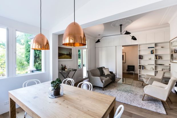 A bright open-plan interior combining a dining area and living room. The foreground features a large light timber dining table with white chairs and pendant lights with copper-toned shades. Beyond it is a refined living area with a grey sofa, white lounge chairs, patterned rug, built-in white shelving, decorative wall panelling, framed artwork, and tall windows overlooking greenery. The space has high ceilings, ornate ceiling detailing, and a calm, light neutral palette.