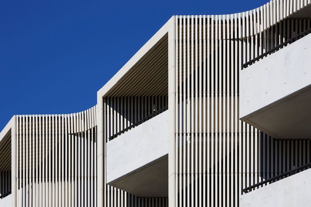 Close-up exterior view of a contemporary multi-storey building with crisp white concrete forms, deep recessed balconies, and extensive vertical white screening slats. The geometric facade creates strong linear shadows against a clear blue sky.