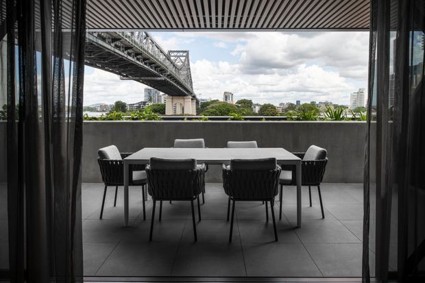 A contemporary outdoor dining terrace viewed from inside through open glazing, with a rectangular dining table and six dark woven chairs on large grey floor tiles. A slatted overhead canopy provides shade, while a solid parapet frames views toward a large steel bridge, riverfront vegetation, and a city skyline beyond.