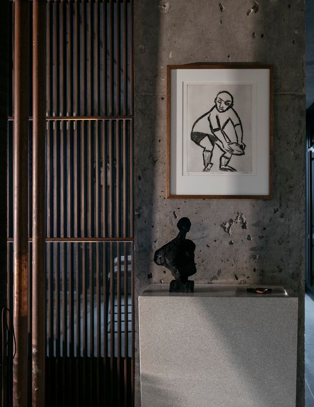 A moody interior vignette featuring a rough exposed concrete wall, a framed monochrome figurative artwork, and a dark sculptural object displayed on a simple pale stone or concrete console. To the left, a tall timber-framed screen of closely spaced dark vertical battens partially separates the space. The composition is minimal, textural, and gallery-like, with strong directional light and deep shadows.