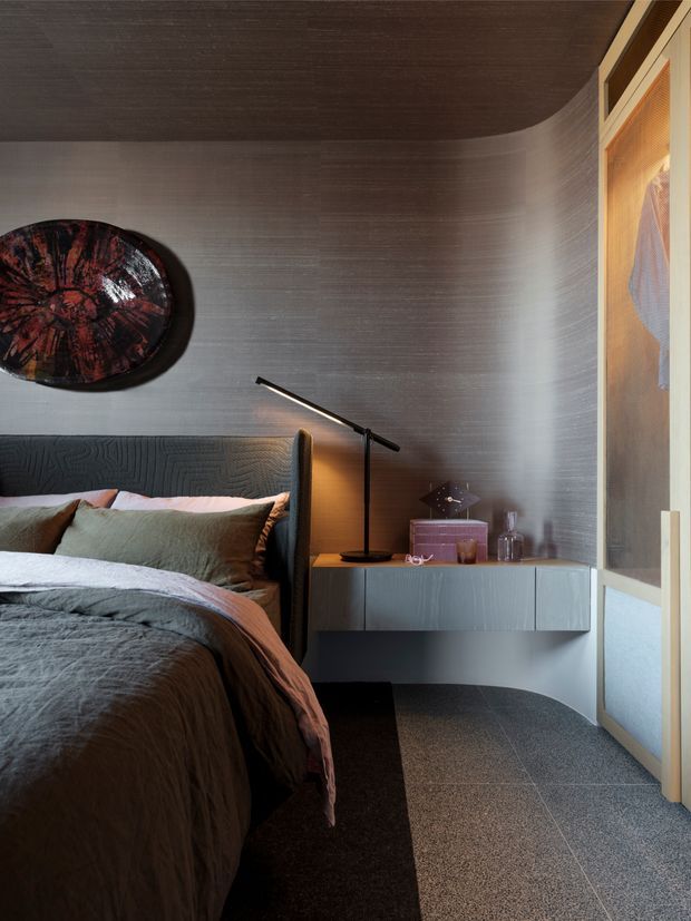 A refined, moody bedroom interior with a partially visible bed, textured charcoal headboard, floating bedside cabinet, slim black table lamp, layered bedding, and dark wall and ceiling finishes. The room features softly curved wall detailing, a dark tiled or stone-like floor, and warm timber-framed textured glazing or artwork at the right.