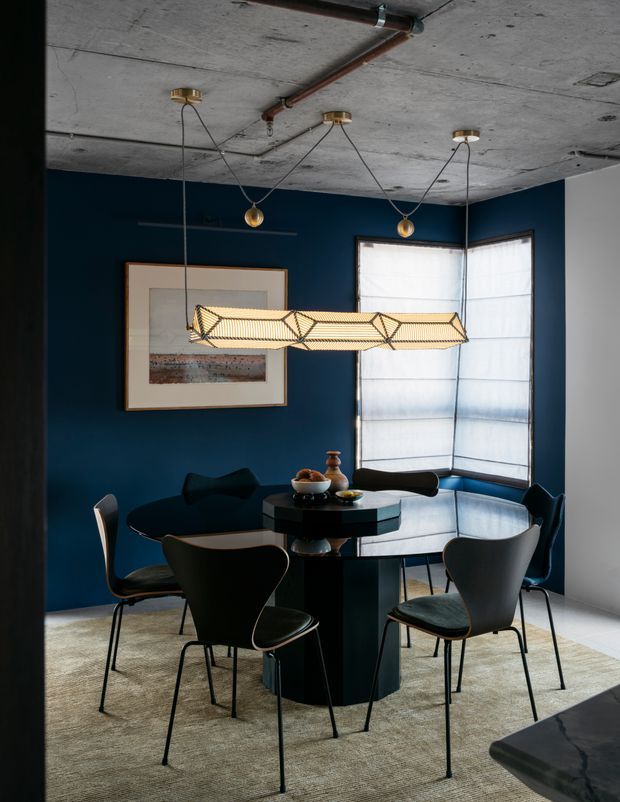 A moody, stylish dining room with a dark navy-blue feature wall, round black dining table, and sculptural black dining chairs arranged beneath a geometric woven pendant light. The room has a raw exposed concrete ceiling with visible copper-toned pipework and suspended globe lights. A large corner window with translucent blinds provides soft daylight, while framed artwork, a neutral textured rug, and minimal table decor complete the space.