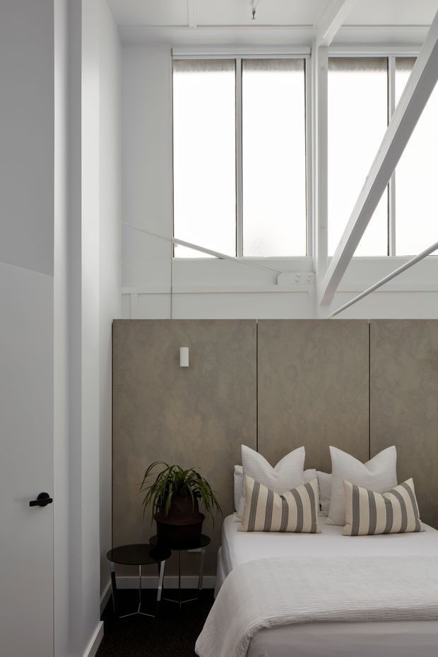 A bright, compact bedroom with a white-painted industrial-style interior, tall frosted windows, exposed structural beams, and a high ceiling. The bed is dressed in white linen with striped neutral cushions and set against a full-height taupe textured headboard. A small black side table and potted plant sit beside the bed, while a white door with a black lever handle is visible in the foreground.