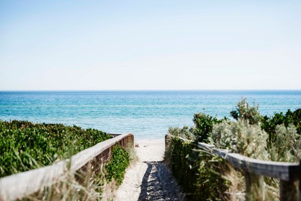 A sandy coastal walkway bordered by low timber railings and dense dune vegetation leads directly toward a bright turquoise ocean and clear horizon.