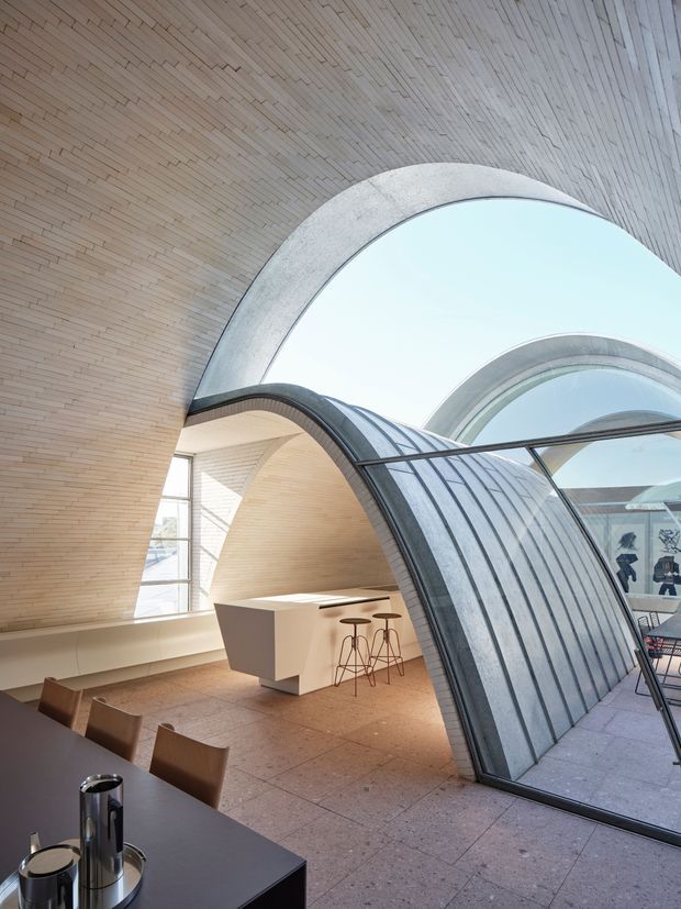 A dramatic contemporary interior with a sculptural vaulted form and large arched glazed roof structure. Pale narrow brick or tile-like lining covers the curved ceiling and walls, while a dark ribbed metal-framed arch encloses a bright atrium-like space. A minimalist white island with bar stools sits beside a dark dining table and timber chairs. The floor appears to be large-format warm grey stone or terrazzo tiles, and daylight fills the space.