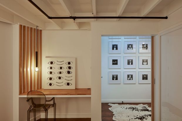 A warm, minimalist interior passage or entry area with white walls and exposed painted ceiling beams. A built-in timber desk or console spans the left wall beneath a vertical timber slat feature, with a transparent chair tucked underneath. A monochromatic artwork rests on the desk, while the opening ahead reveals a display wall of framed photographs, dark timber flooring, and a cowhide rug. Black exposed services piping runs across the ceiling, adding a subtle industrial detail.