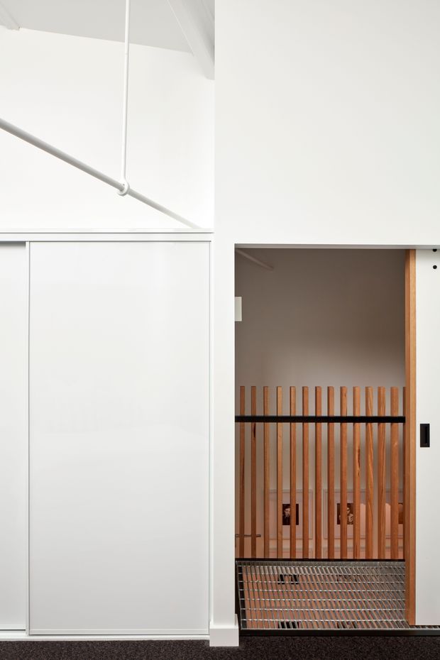 A minimalist interior view of a stairwell or landing, featuring crisp white walls and concealed white cabinetry or sliding panels. An open stair opening is enclosed by vertical timber balusters with black horizontal rails, revealing a metal mesh stair tread or landing below. The ceiling includes exposed white conduit or pipework, and the space is finished with dark carpet flooring.