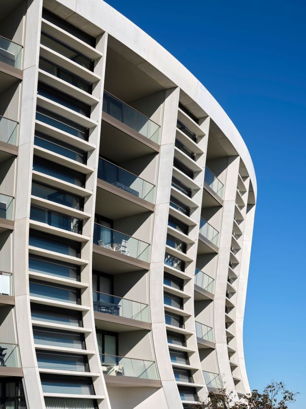Low-angle exterior photograph of a modern multi-storey apartment building with a distinctive curved facade. Repetitive recessed balconies, pale concrete or rendered structural framing, dark glazed windows, and transparent glass balustrades create a strong geometric pattern against a clear blue sky.