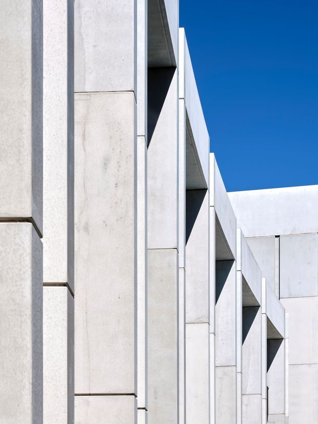 Abstract close-up of a stark geometric building facade composed of pale concrete panels and deep recessed vertical openings. Repeating projecting and recessed elements create a strong rhythm and dramatic shadow pattern against a clear blue sky. The image focuses on the exterior envelope with no visible interior spaces or furnishings.