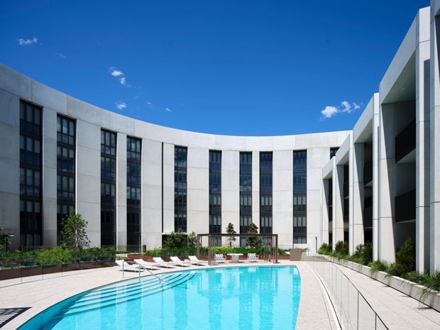 A bright architectural exterior courtyard with a large turquoise swimming pool enclosed by a sweeping curved, multi-storey modern building. The facade is composed of pale concrete or rendered panels, tall dark-framed windows, deep recessed openings, and strong geometric vertical elements. A paved pool terrace with lounge chairs, low planting beds, and a pergola-like structure occupies the foreground under a clear blue sky.