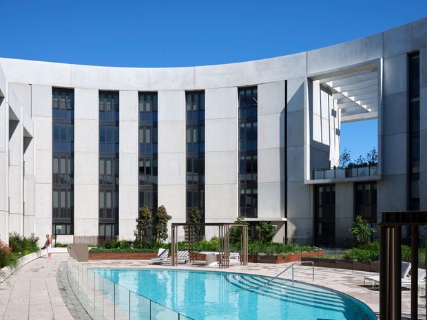 A bright exterior view of a contemporary multi-storey building arranged around a landscaped courtyard and swimming pool. The curved facade uses pale concrete or stone-like panels with tall dark-framed glazing, while a deep rectangular opening and projecting pergola-like structure create a strong architectural focal point. The pool, paved terrace, planting, and outdoor seating occupy the foreground under a clear blue sky.
